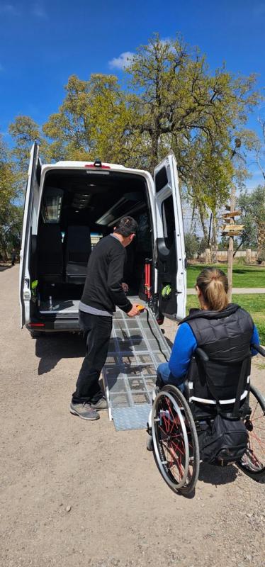A woman using a wheelchair beside an adapted van while an assistant prepares the access ramp in Mendoza.